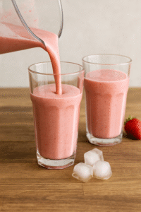 Pouring strawberry smoothie into tall glasses on a wooden table.