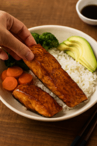 Assembling Teriyaki Salmon Bowl with rice, glazed salmon, broccoli, carrots, and avocado slices on a wooden countertop.