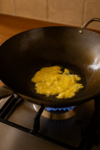 Soft scrambled eggs cooking in sesame oil inside a wok as the first step for Vegetable Fried Rice, with steam rising and golden texture visible.