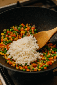 Cooked white rice added into a wok with vegetables while preparing Vegetable Fried Rice, mixed with a wooden spatula under natural kitchen lighting.