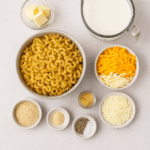 Mac and Cheese ingredients – macaroni pasta, butter, flour, milk, cheddar, mozzarella, Parmesan, breadcrumbs, and seasonings on table.