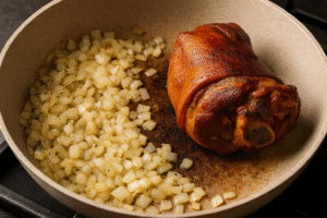 Varză călită cu ciolan afumat – smoked pork hock and sautéed onions sizzling in oil inside a rustic pan.