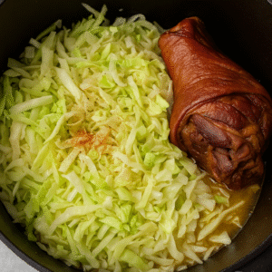 Varză călită cu ciolan afumat – fresh cabbage added to the pot with smoked pork hock and onions, starting to soften.