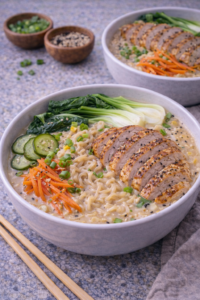 Bowl of creamy sesame chicken ramen topped with sliced chicken, bok choy, carrots, cucumber, green onions, and sesame seeds.