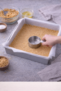 Pressing graham cracker crust into a lined baking pan for strawberry cheesecake bars.